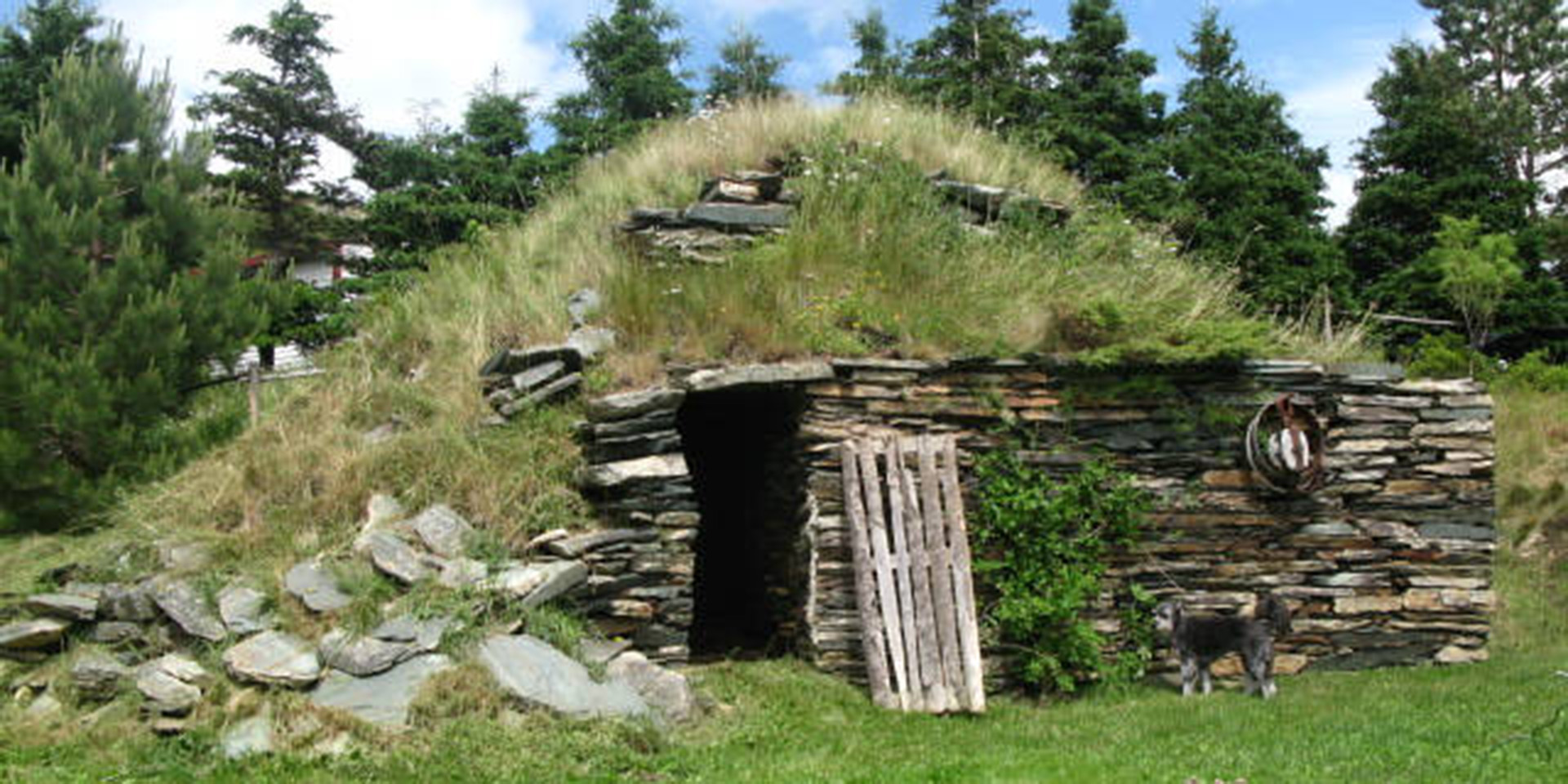 Extérieur d’une cave à légumes à flanc de colline, avec façade en pierres empilées et dessus recouvert d’herbe.