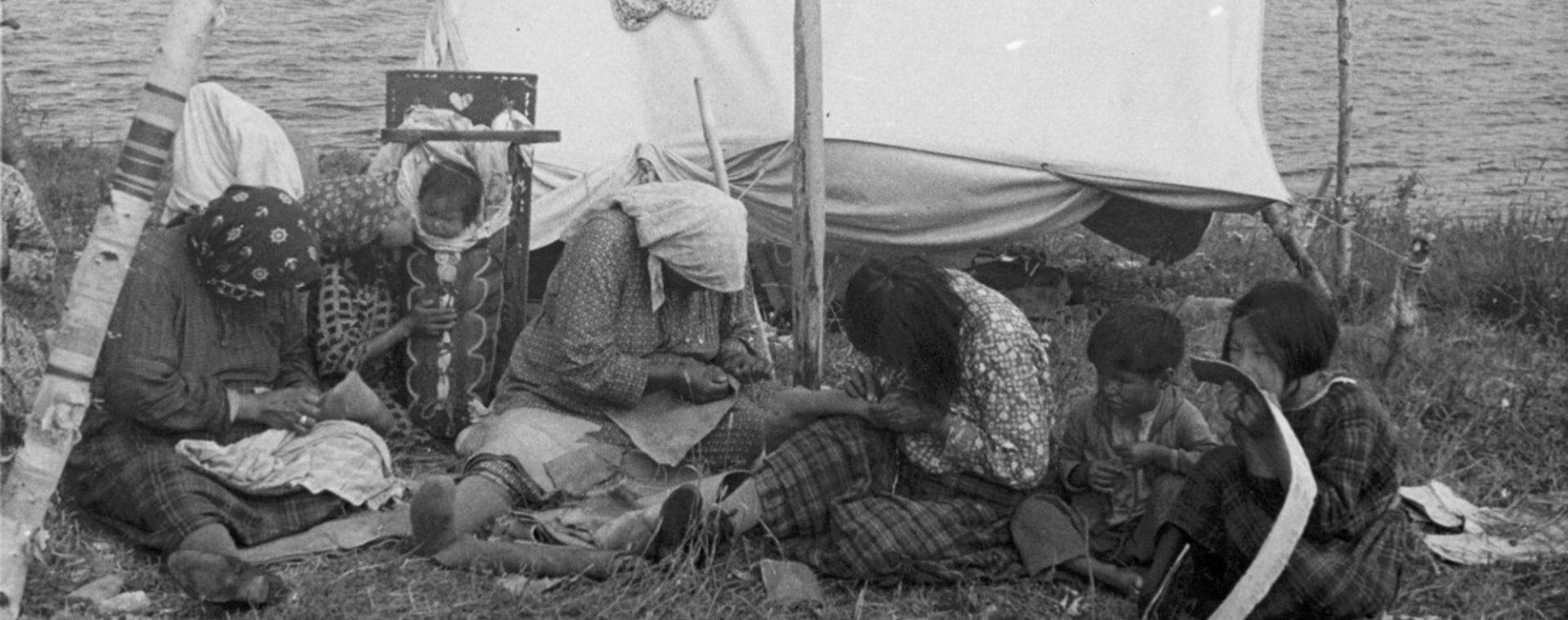 La photo est en noir et blanc. Des femmes et des enfants anicinabek sont assis dans l’herbe près du lac qu’on aperçoit en arrière-plan. Ils sont près d’une petite tente en toile et les femmes s’affairent à la couture de mocassins alors que les enfants les observent ou jouent. Un bébé est installé dans un tikinagan, un porte-bébé traditionnel.