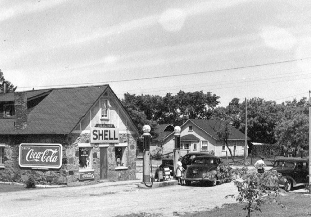 Photo en noir et blanc de voitures anciennes stationnées devant les pompes à essence avec l’édifice en pierre à l’arrière-plan