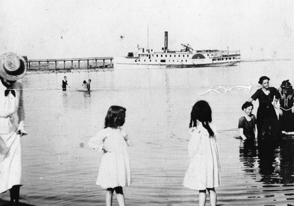 Photographie en noir et blanc d'un lac avec des individus debout dans l'eau et à l'arrière-plan un grand bateau à vapeur et une jetée