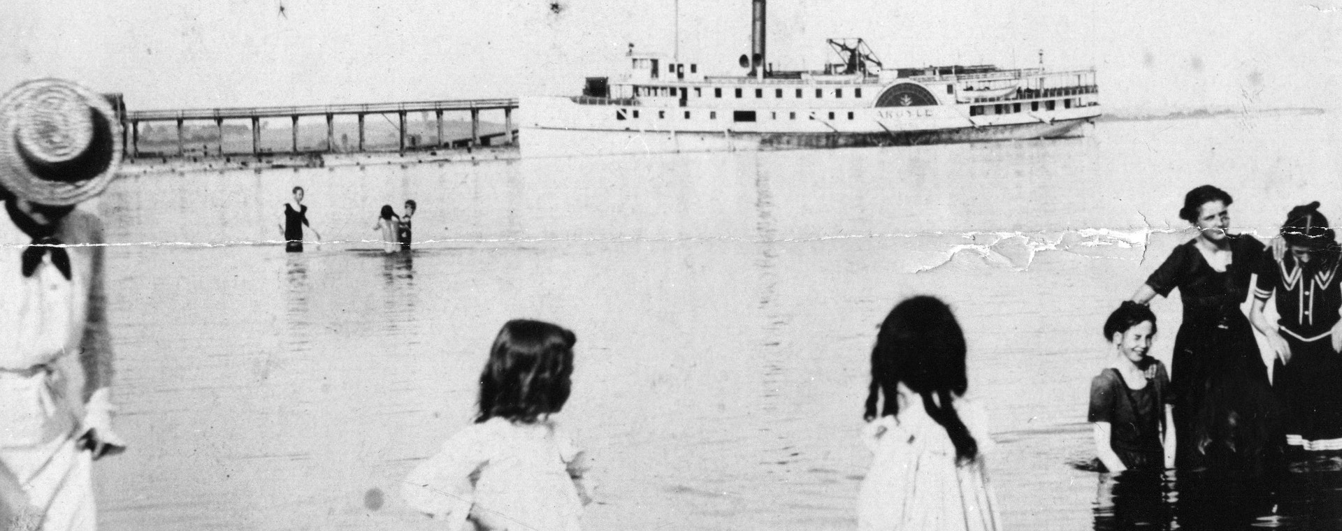 Photographie en noir et blanc d'un lac avec des individus debout dans l'eau et à l'arrière-plan un grand bateau à vapeur et une jetée