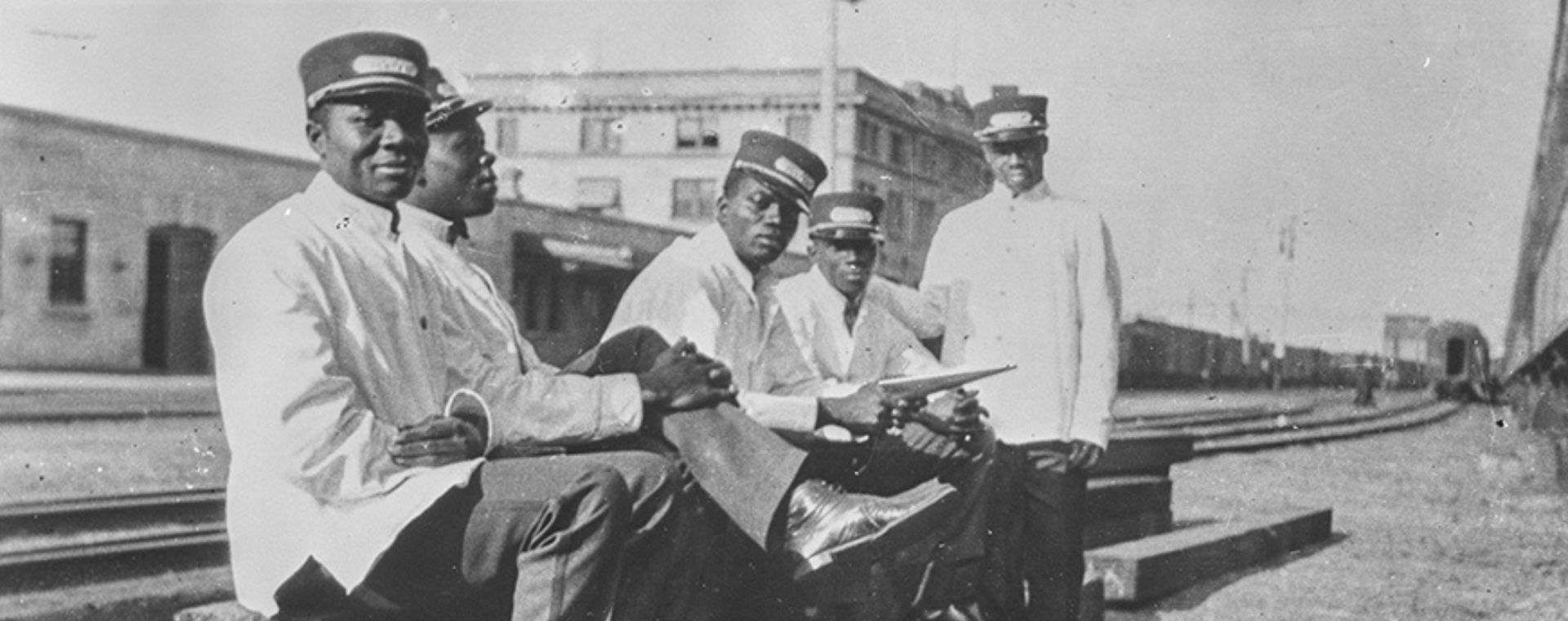 Photographie d’archives en noir et blanc de cinq hommes noirs assis sur une voie ferrée. Ils sont vêtus d’uniformes ferroviaires blancs.