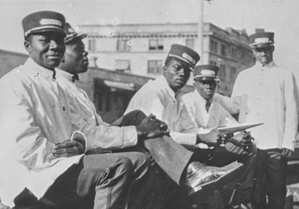 Photographie d’archives en noir et blanc de cinq hommes noirs assis sur une voie ferrée. Ils sont vêtus d’uniformes ferroviaires blancs.