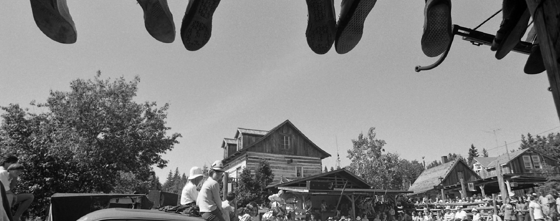 Photo noir et blanc d’une centaine de spectateurs assistant à une prestation extérieure à La Butte. Le haut de l’image montre les pieds de personnes installées sur une partie de la toiture de la Butte.