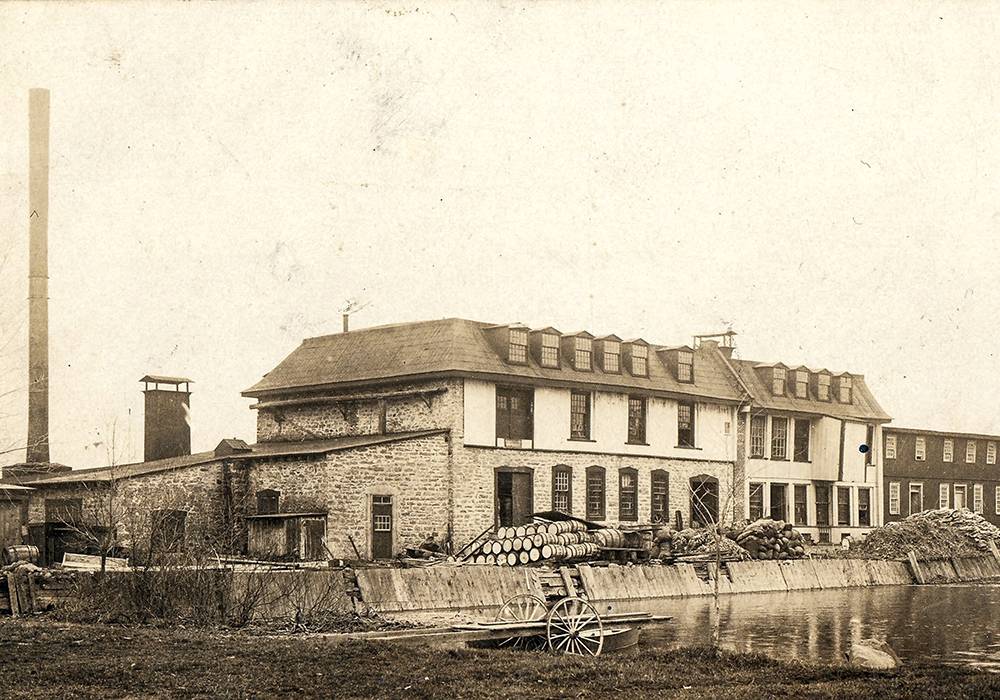 Carte postale publiée vers 1900. Au centre : la fabrique de papier avec, à sa gauche, sa haute cheminée sur la digue des moulins encombrée de tonneaux et de matériaux. À l'avant-plan: un quai roulant donne sur le bassin d’eau.