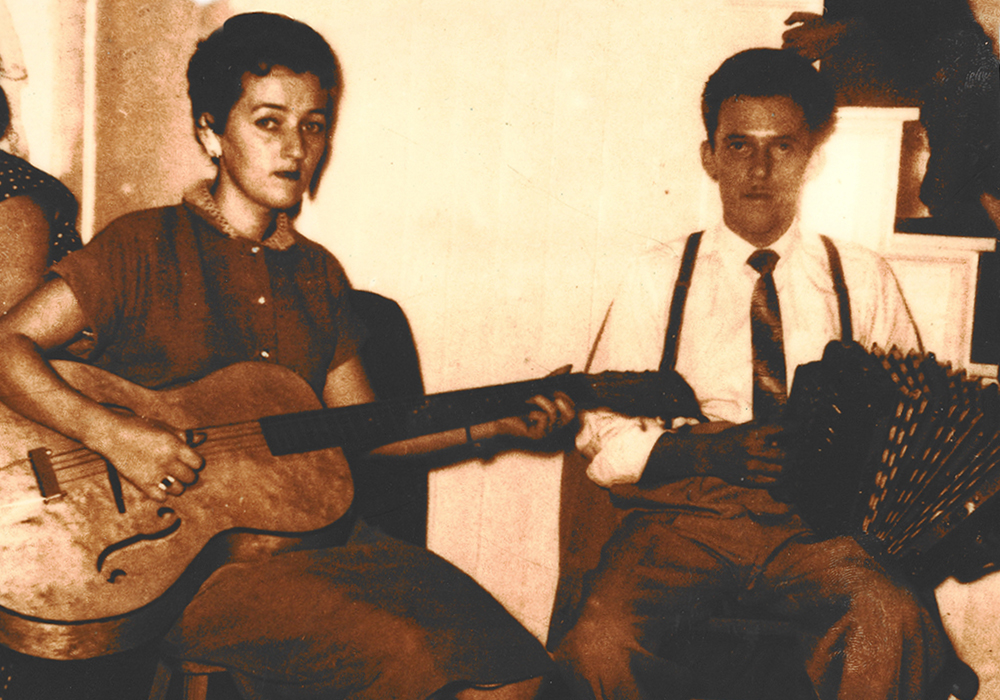 Yvette Coulombe est assise à la gauche d’Ulric Lacombe avec une guitare sur ses genoux. Ulric Lacombe est assis à sa droite avec un accordéon sur ses genoux.
