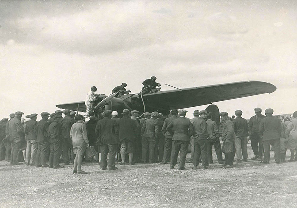 Photo en noir et blanc de trois personnes se trouvant sur les ailes d’un avion, lors du ravitaillement. Une foule nombreuse se tient autour de l’avion, dos à l’appareil-photo.