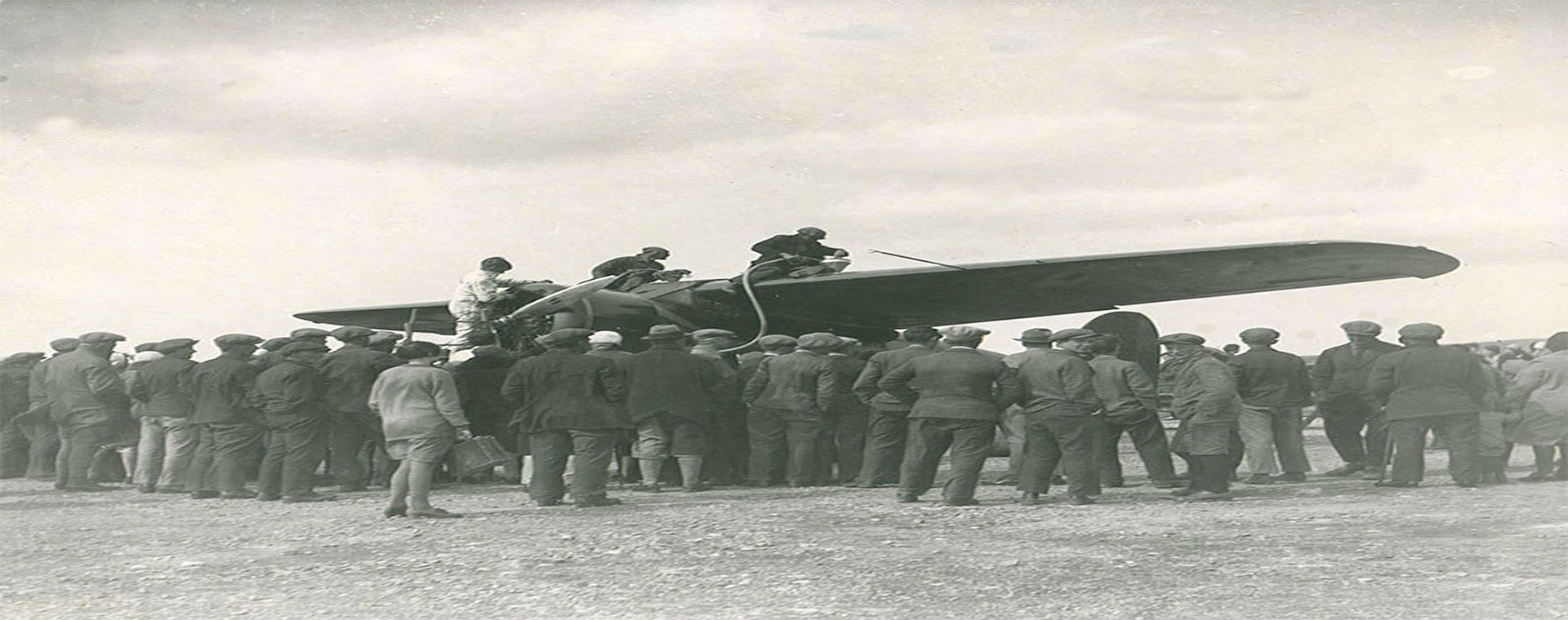 Photo en noir et blanc de trois personnes se trouvant sur les ailes d’un avion, lors du ravitaillement. Une foule nombreuse se tient autour de l’avion, dos à l’appareil-photo.