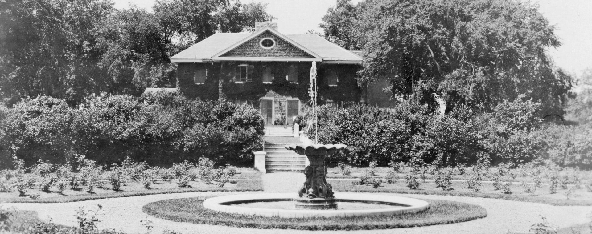 Photographie en noir et blanc d’un jardin circulaire d’arbustes entourant une fontaine devant une grande maison en pierre de deux étages, couverte de plantes grimpantes et munie d’un escalier donnant sur le jardin