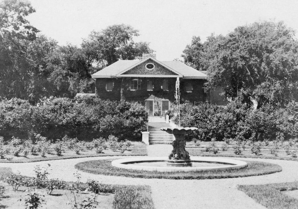 Photographie en noir et blanc d’un jardin circulaire d’arbustes entourant une fontaine devant une grande maison en pierre de deux étages, couverte de plantes grimpantes et munie d’un escalier donnant sur le jardin