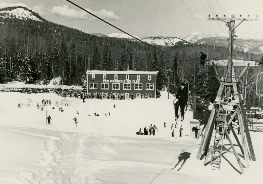 Photo en noir et blanc d’un skieur sur un télésiège. Au loin, on voit le chalet du Club de Ski de Red Mountain