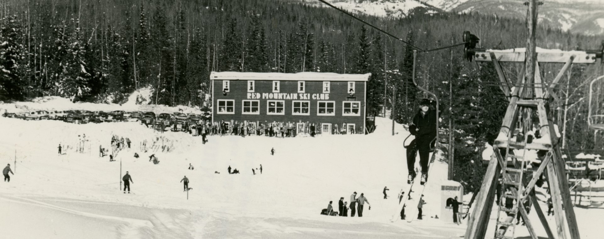 Photo en noir et blanc d’un skieur sur un télésiège. Au loin, on voit le chalet du Club de Ski de Red Mountain