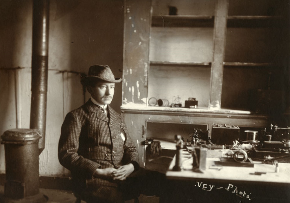 Un homme vêtu d'un veston à carreaux et d'un chapeau est assis à une table sur laquelle est posé du matériel de communication sans fil. Derrière lui se trouvent une armoire vide sans portes, ainsi qu’un poêle et un tuyau de poêle en métal.