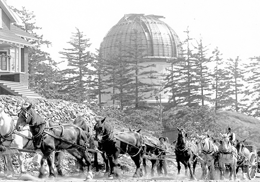 A black and white photograph showing part of a 1.8 m telescope being hauled by horse-drawn wagon, with telescope dome and the Director’s residence in background