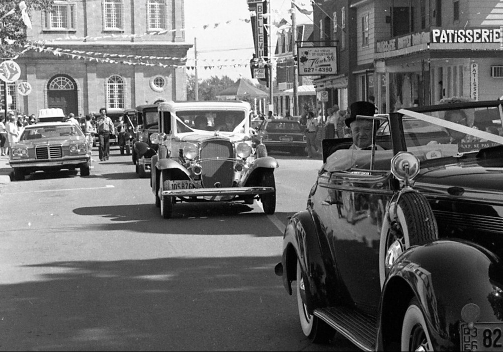 Photographie en noir et blanc de voitures défilant sur la rue Saint-Eustache. Des commerces sont visibles sur le côté droit de la rue et l'église Saint-Eustache est en arrière-plan.