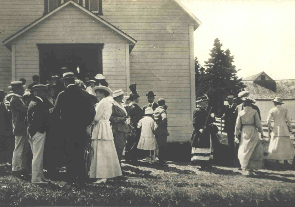 Photographie noir et blanc d'hommes et de femmes attroupés devant un église de bois à la fin de la messe. Les hommes sont, en majorité, vêtu d'un habit au veston foncé, d'un pentalon blanc et d'un chapeau tressé plat décoré d'un ruban. Les femmes portent une longue robe blanche et un chapeau plat tressé. En arrière plan, on aperçoit derrière un massif d'épinettes matures, un bâtiment de ferme.