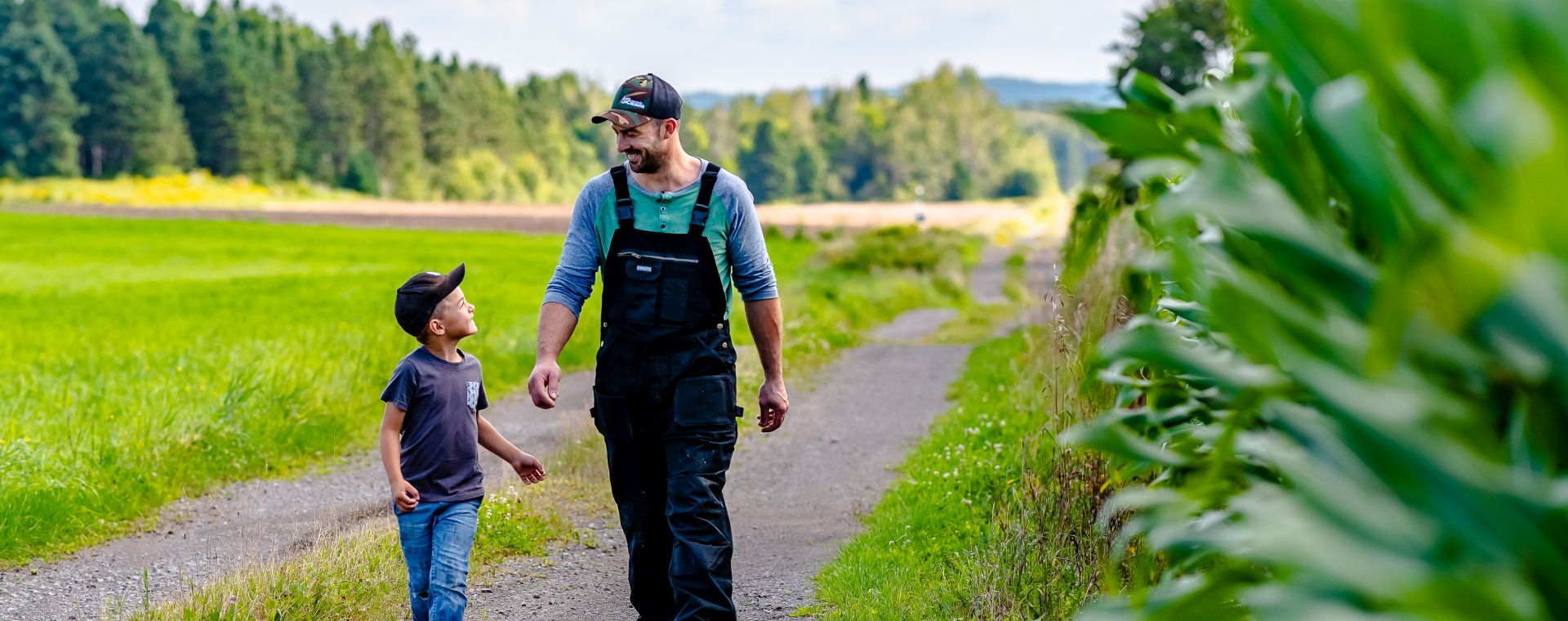 Un agriculteur en salopette et casquette marche côte à côte avec un jeune garçon sur un sentier de terre. Ils sont au milieu de champs cultivés. Les deux sourient, devant une rangée d'arbres et un ciel bleu nuageux.