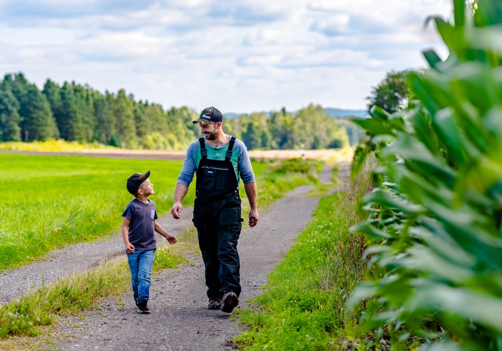 Un agriculteur en salopette et casquette marche côte à côte avec un jeune garçon sur un sentier de terre. Ils sont au milieu de champs cultivés. Les deux sourient, devant une rangée d'arbres et un ciel bleu nuageux.