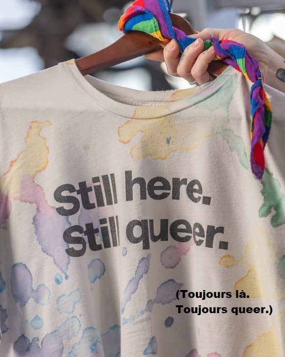 Photo d’un bras tatoué tenant un t-shirt blanc cassé sur un cintre en bois. Le t-shirt comporte un motif multicolore de taches aléatoires et sans formes ressemblant à de l’encre ou à de l’aquarelle. Sur le devant, le texte en noir en anglais se traduit « Toujours là. Toujours queer. ». Un mouchoir à rayures arc-en-ciel est noué en haut du cintre.