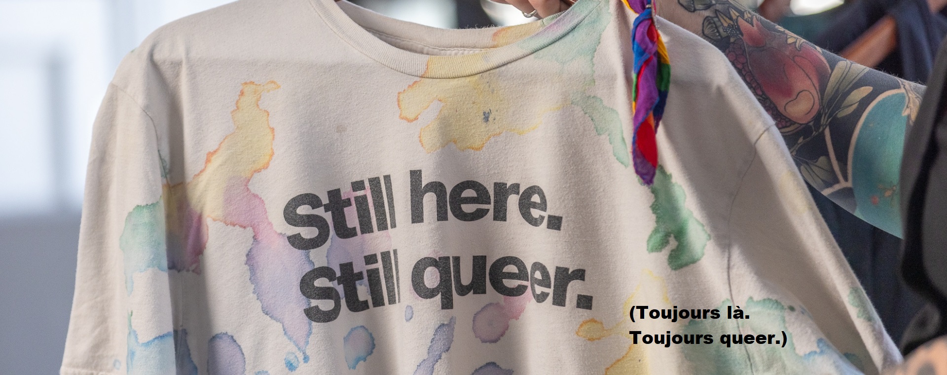 Photo d’un bras tatoué tenant un t-shirt blanc cassé sur un cintre en bois. Le t-shirt comporte un motif multicolore de taches aléatoires et sans formes ressemblant à de l’encre ou à de l’aquarelle. Sur le devant, le texte en noir en anglais se traduit « Toujours là. Toujours queer. ». Un mouchoir à rayures arc-en-ciel est noué en haut du cintre.