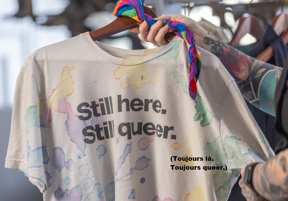 Photo d’un bras tatoué tenant un t-shirt blanc cassé sur un cintre en bois. Le t-shirt comporte un motif multicolore de taches aléatoires et sans formes ressemblant à de l’encre ou à de l’aquarelle. Sur le devant, le texte en noir en anglais se traduit « Toujours là. Toujours queer. ». Un mouchoir à rayures arc-en-ciel est noué en haut du cintre.